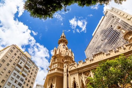 Edifício histórico de estilo gótico, com torre central pontiaguda e ornamentos detalhados, visto de baixo contra um céu azul com nuvens. Ao redor, há prédios modernos e árvores na parte superior e inferior da imagem.