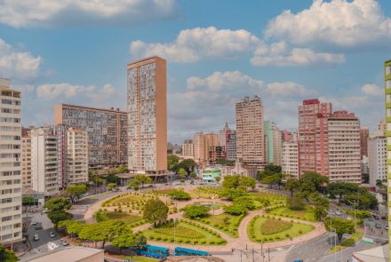 Vista aérea diretamente de cima, mostrando o desenho simétrico da Praça Raul Soares. Os caminhos claros formam padrões geométricos que se cruzam. No centro, a fonte redonda tem água de cor verde. Os espaços são preenchidos por grama, arbustos esféricos e algumas árvores. Pessoas caminham no local.
