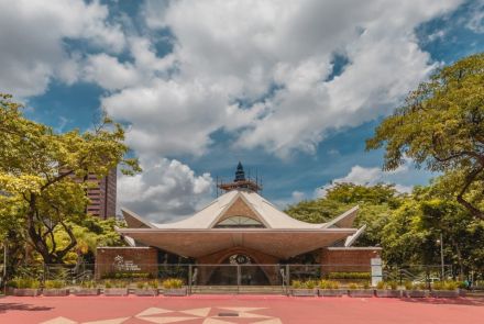 Vista frontal de igreja com telhado branco geométrico em abas inclinadas. No topo, torre azul com cruz. A fachada combina tijolos aparentes e vitrais triangulares. Em frente, canteiros baixos com plantas e piso em vermelho e bege. Árvores grandes cercam o espaço.