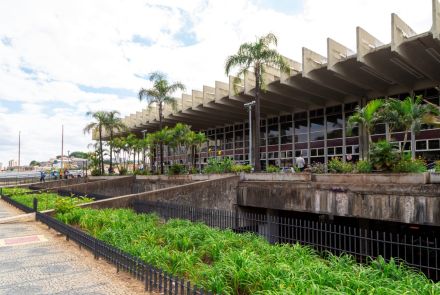 Vista lateral externa do edifício da rodoviária. A marquise de concreto, com seu design de vigas em série, cobre a fachada de vidro. Em frente ao prédio, há um jardim em desnível com palmeiras e vegetação baixa, separado da calçada por uma mureta de concreto e uma grade de metal preta.