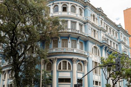 Edifício azul de esquina, de vários andares, com colunas brancas na fachada, sacadas com grades metálicas e cúpula arredondada no topo. Pessoas passam pela calçada, e há lojas no nível da rua.