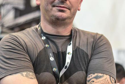 Foto de um homem posando para foto de braços cruzados, esboçando um sorriso de boca fechada. O homem é branco careca e sem barba. O homem usa blusa cinza de mangas curtas, relógio marrom no pulso direito e tem nos braços tatuagens na cor preta. 