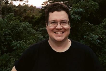 Foto de um homem em pé, ao ar livre, aparecendo da cintura para cima, com várias árvores ao fundo. O homem posa para a foto sorrindo. O. homem tem cabelos castanhos escuros, usa óculos de grau e veste blusa preta de mangas curtas.