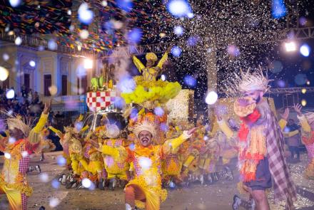 Fotografia da apresentação da Quadrilha Luar de São João. À frente da imagem temos pedaços de confete desfocados. No plano principal da foto, os integrantes da quadrilha dançam no tablado com vestimentas amarelas remendadas por panos vermelhos quadriculados, eles estão de joelhos e com os braços levantados para o céu rodeando um carro alegórico, que por sua vez destaca uma dançarina que utiliza vestido amarelo com paetê. Ao fundo da imagem está uma parte do prédio histórico da praça da estação.