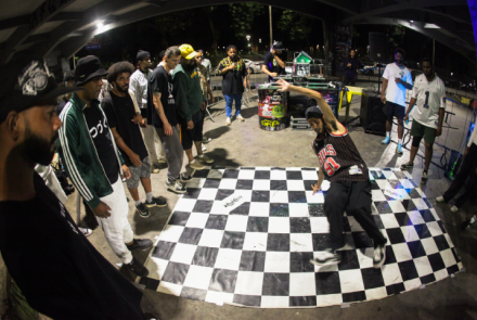 A imagem mostra um grupo de pessoas em um ambiente urbano, participando de uma competição ou batalha de dança em estilo hip-hop. No centro, um dançarino está realizando um movimento dinâmico sobre um tablado quadriculado, enquanto os outros participantes, em pé ao redor, assistem com interesse. O local parece iluminado por luzes artificiais, com uma mesa de DJ ao fundo e um clima animado de confraternização e arte de rua.