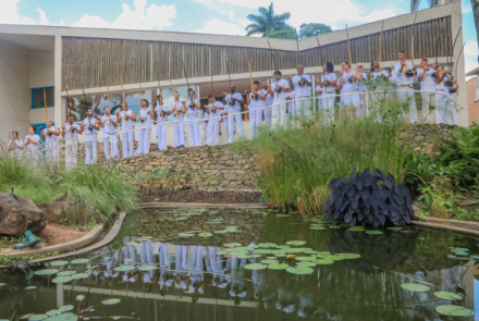 Na imagem um grupo de capoeira está vestido de branco tocando berimbau em frente à varanda do museu casa kubitschek. a foto enquadra em primeiro plano o lago do museu e em segundo plano os integrantes do grupo oficina de capoeira em linha reta tendo ao fundo o telhado borboleta da edificação.
