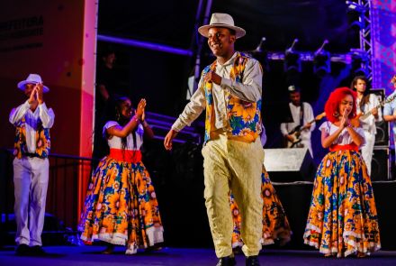 A imagem mostra um grupo de pessoas em um palco durante uma apresentação cultural vibrante e alegre. Um homem ao centro está sorrindo e dançando, vestindo um colete colorido estampado com girassóis e um chapéu branco, combinando com calças claras. Ele está cercado por outras pessoas também trajadas com roupas tradicionais. Ao fundo, músicos tocam ao vivo, dando energia à cena. O clima é de celebração e representa uma expressão rica da cultura popular brasileira.