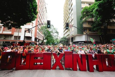 A imagem mostra o bloco de carnaval desfilando por uma rua arborizada, cercada por prédios residenciais, em Belo Horizonte. A imagem mostra um grupo de pessoas segura grandes letras vermelhas tridimensionais que formam a palavra "QUEIXINHO". Também mostra uma multidão animada, vestindo roupas coloridas, adereços carnavalescos e fantasias. O ambiente é vibrante, com muitas pessoas usando acessórios como coroas de flores, óculos escuros e chapéus. A cena transmite alegria, diversidade e a energia contagiante.