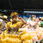 Um grupo de quadrilha junina se apresentando em um tablado, com destaque para uma mulher de vestido amarelo a frente da foto.